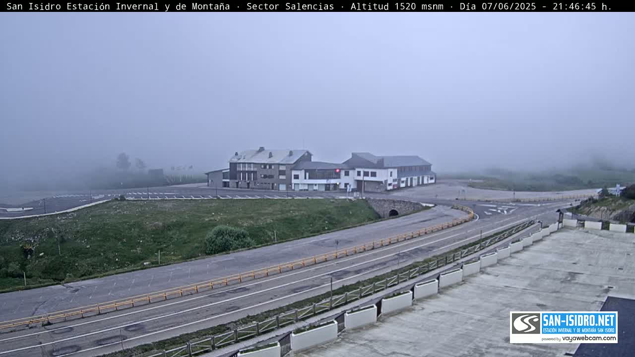 A foggy view of a mountain road curving past a parking lot and two buildings.