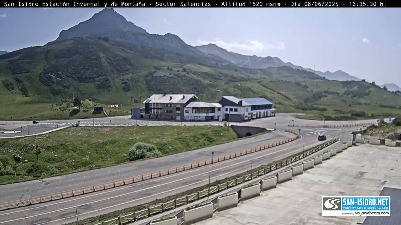 A mountaintop roadway curves past a parking lot and buildings, under a mostly clear sky.