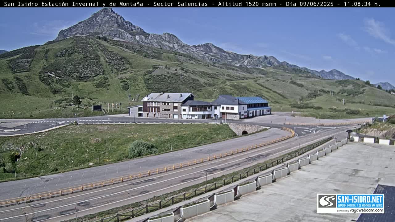 A mountainside scene under sunny skies shows a parking lot, roadway, and several buildings near the base of a green hill.