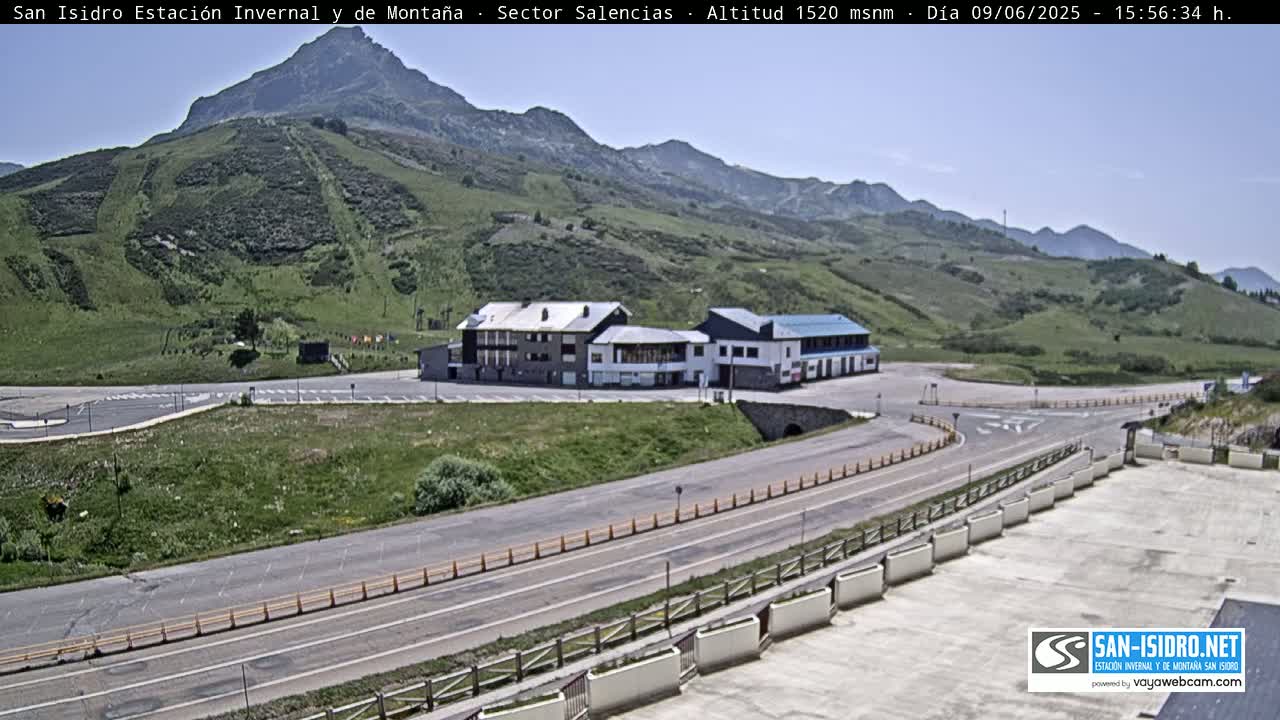 A mountainside resort with a paved road and parking lot is visible under a clear, sunny sky.