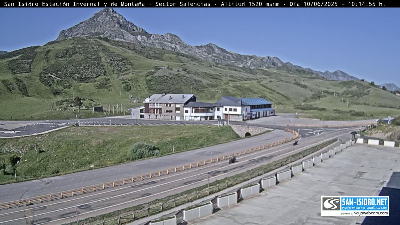 A mountaintop building complex with a parking lot is visible, alongside a road with a few vehicles, all under a clear, sunny sky.