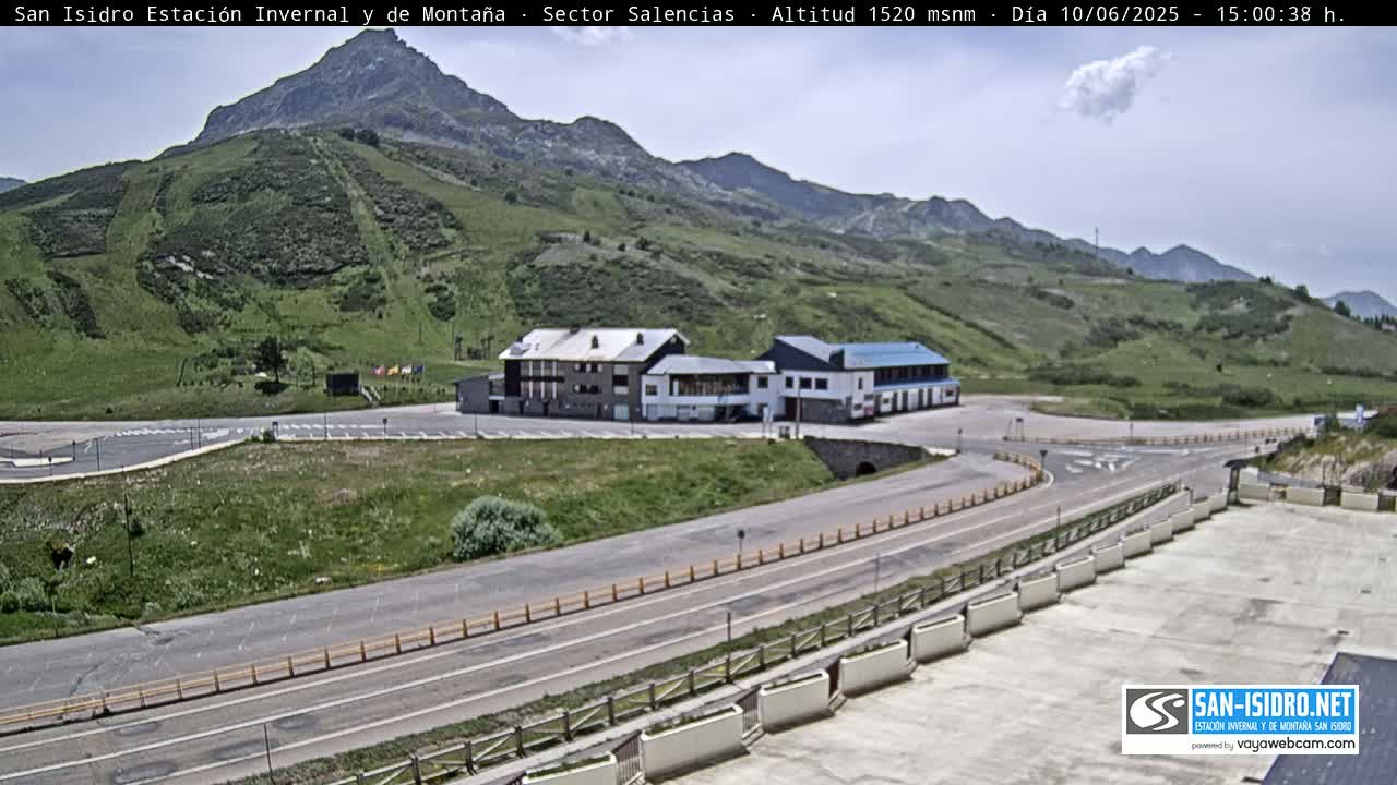 A mountain resort with a building complex and paved roads is situated on a green hillside under a mostly clear sky.