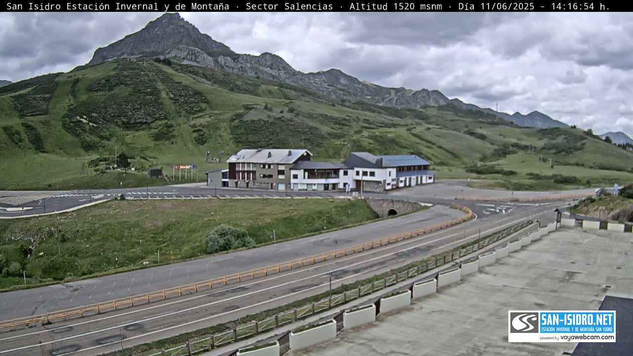 A mountainside resort with several buildings and parking lots is visible under a cloudy sky, with a road curving through the landscape.