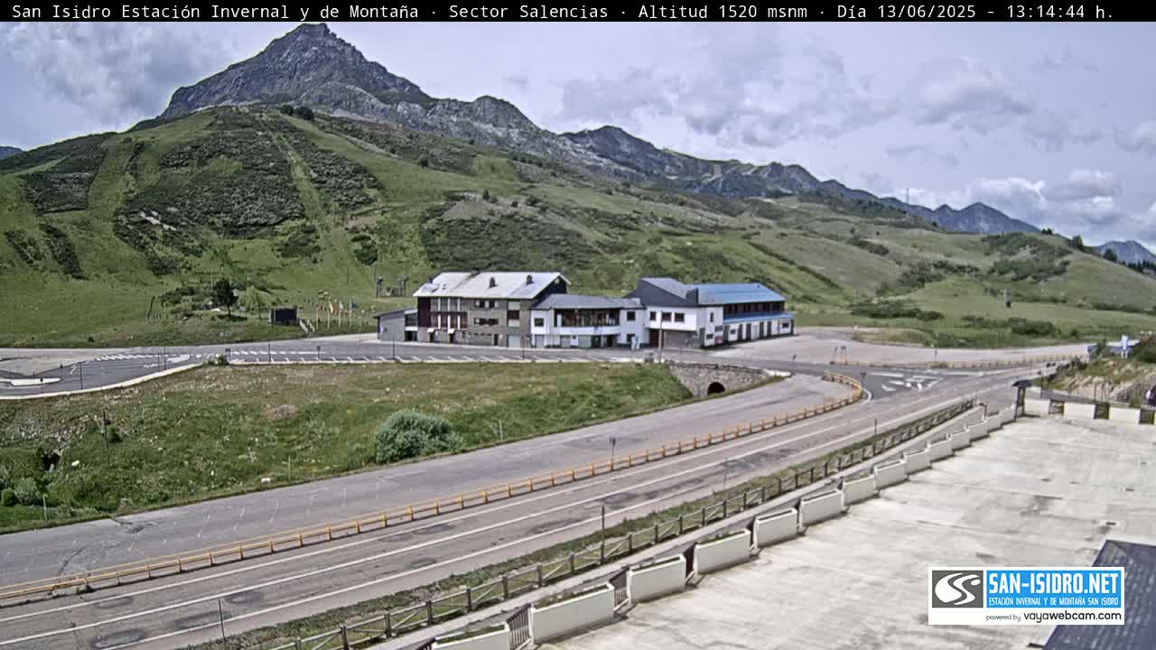 A mountainside parking lot with a building complex sits below a grassy mountain under a partly cloudy sky.