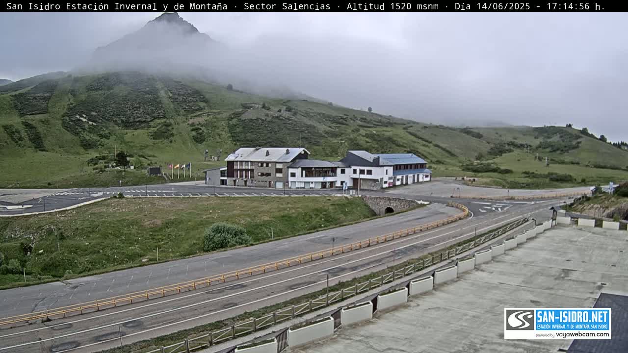 A mountainside with low clouds partially obscuring a mountain peak, overlooks a roadway and buildings in a parking lot.