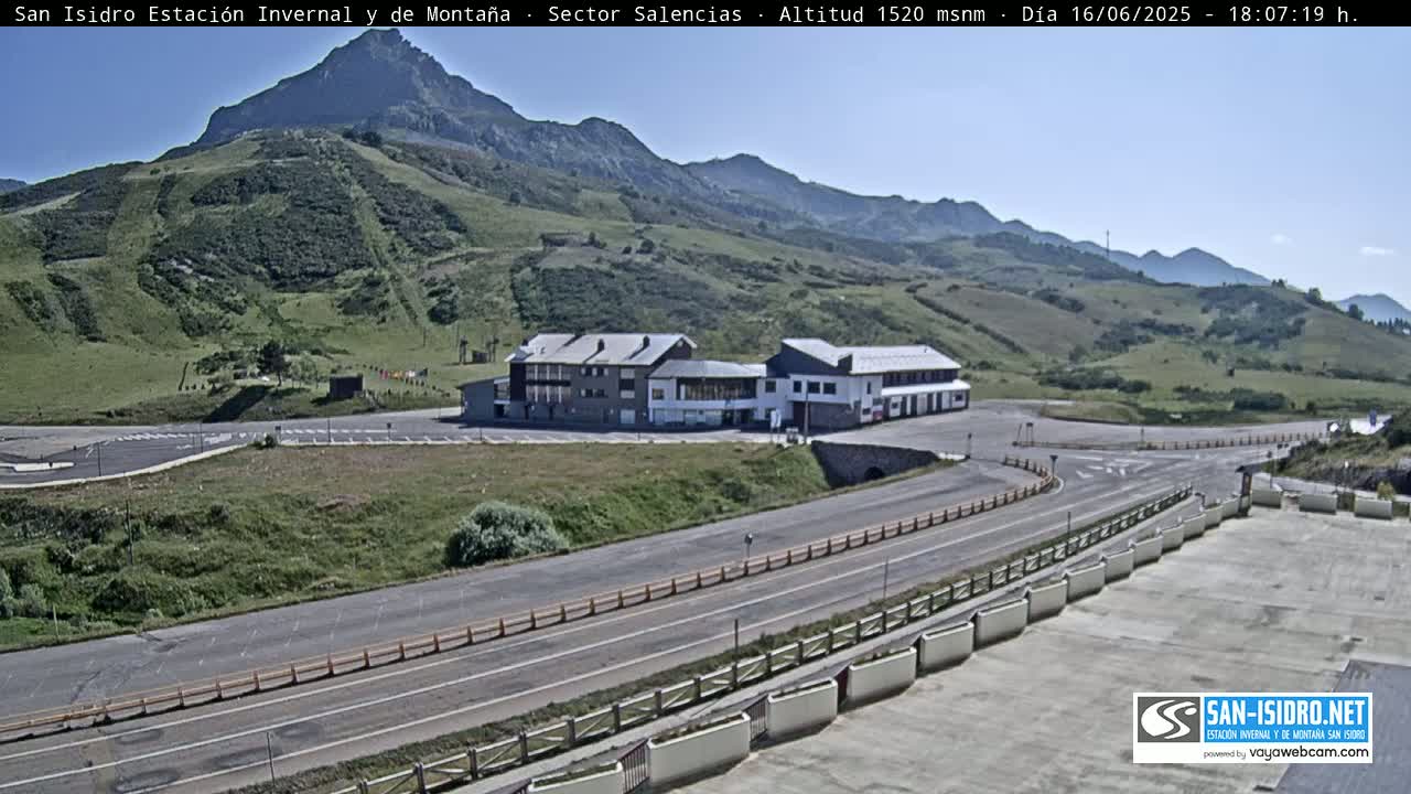 A mountainside building complex with a parking lot and roads sits below a mountain range under a clear sky.