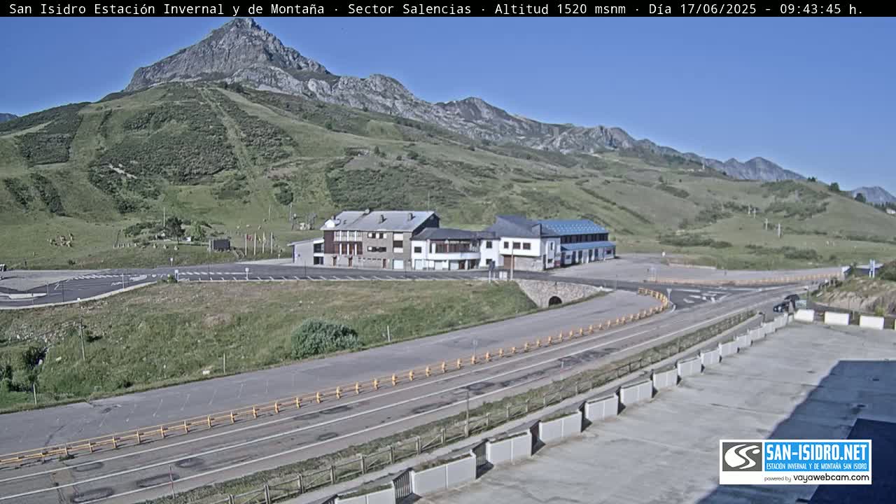 A mountainside resort area with several buildings and a parking lot is visible under a clear blue sky.
