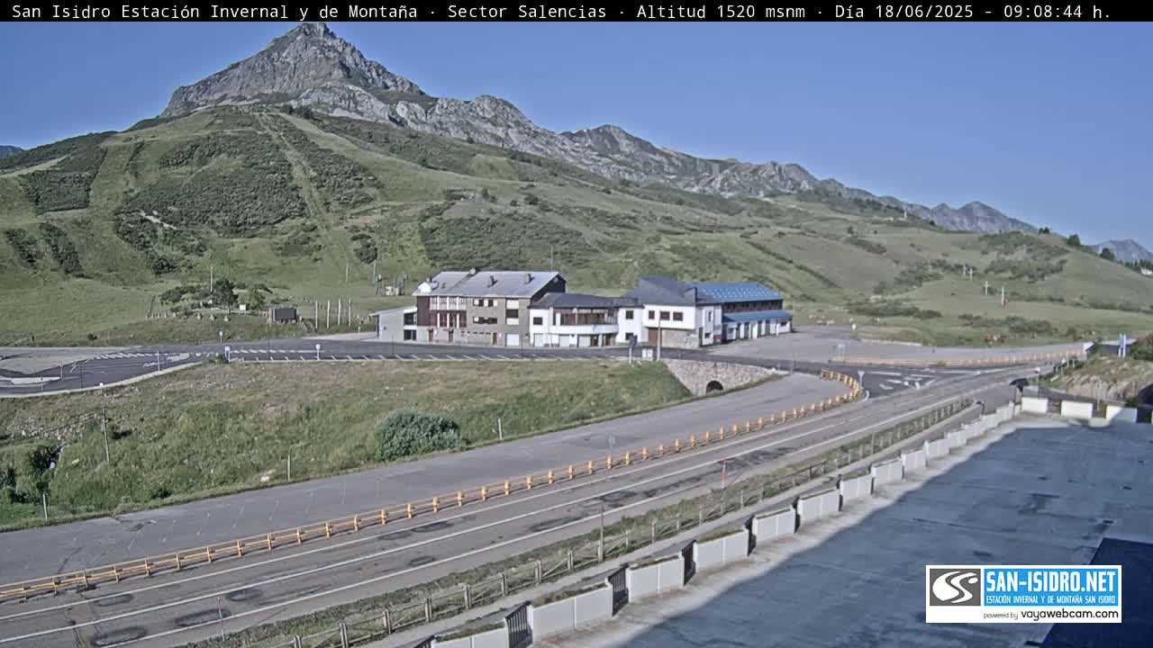 A mountainside parking lot with several buildings sits beneath a clear, sunny sky, with a road curving away from the scene.