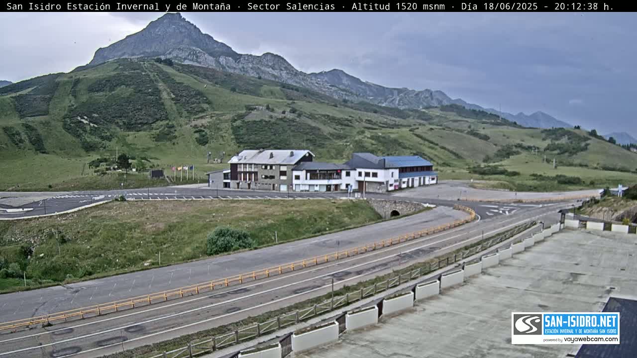 A mountainside parking lot with a few buildings sits below a green hill and a mountain range under an overcast sky.
