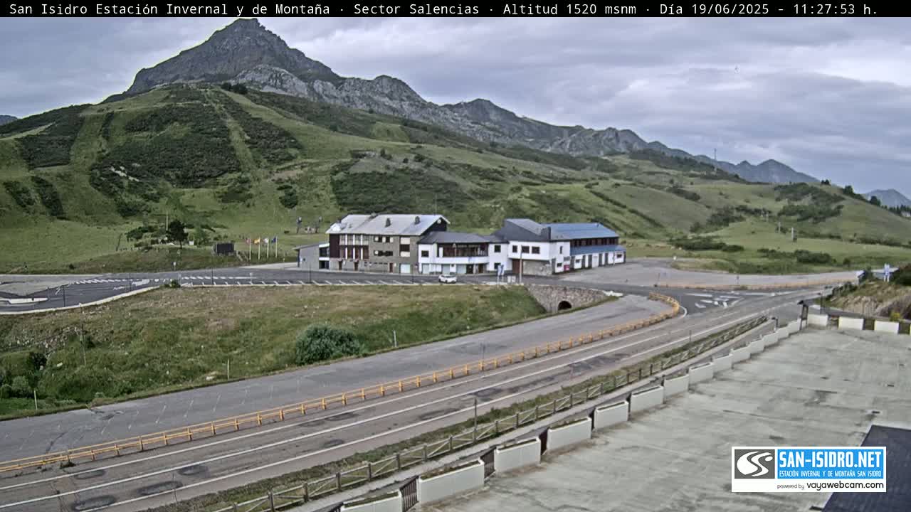 A mountainside view shows a complex of buildings near a roadway curving around a parking lot under a mostly cloudy sky.