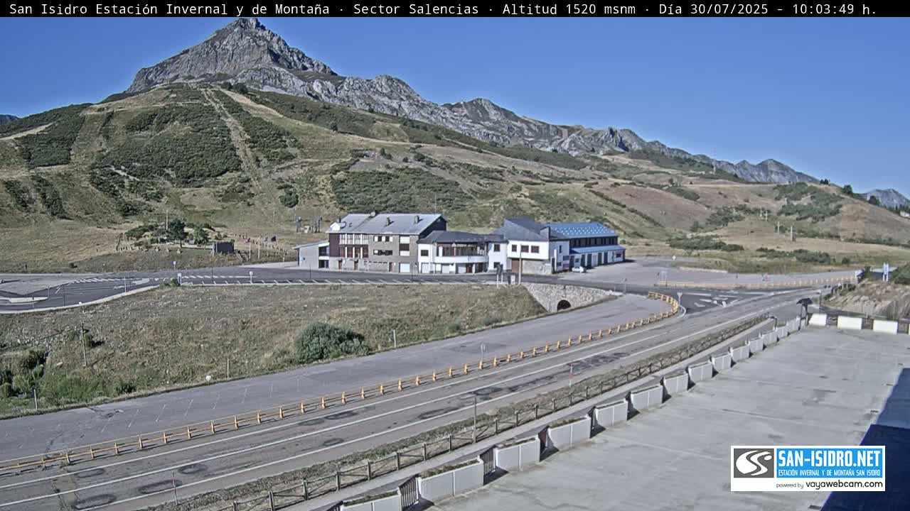 A mountainside parking lot with several buildings is visible on a sunny day, with a road curving around them and leading towards a mountain in the background.
