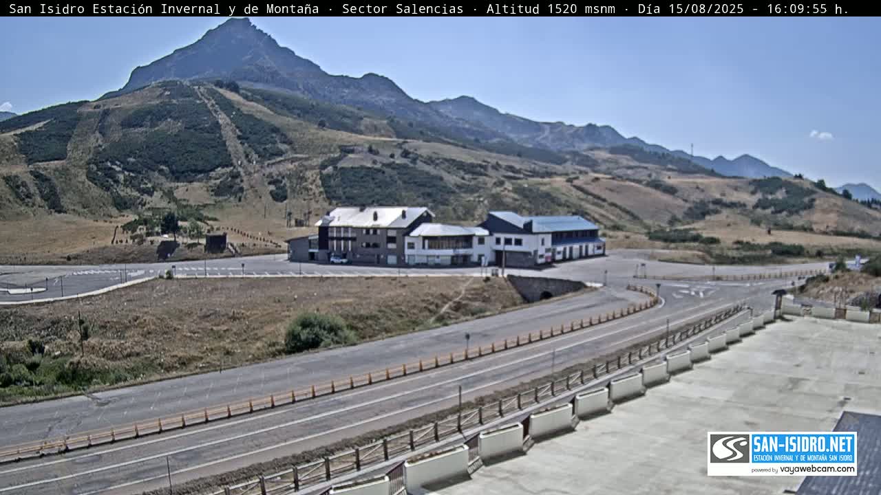 A mountainside parking lot with several buildings is visible on a sunny day, with a road curving around them and leading towards a mountain in the background.