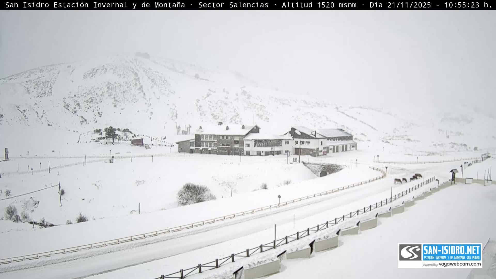 A vast snowy mountain landscape features a multi-building resort complex, a winding road, a small group of horses, and a person with an umbrella, all under heavy snowfall and overcast skies.