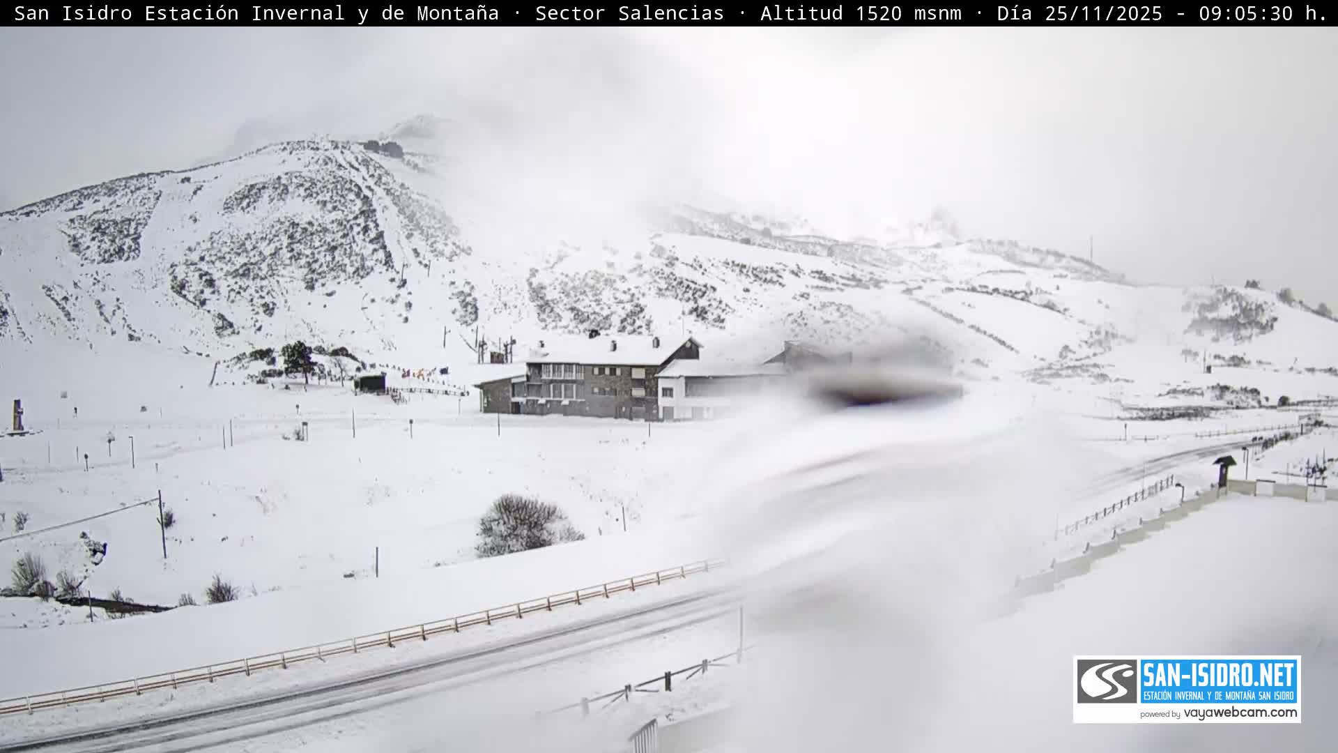 A vast, snow-covered mountain landscape, likely a ski resort, features a large lodge and roads under an overcast, hazy sky with limited visibility.