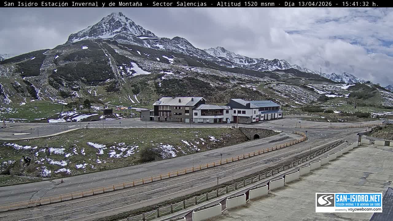 The image depicts a snow-covered mountain ski resort scene with multiple buildings, a partially cleared road with two snowplows, all under heavy snowfall and an overcast sky.