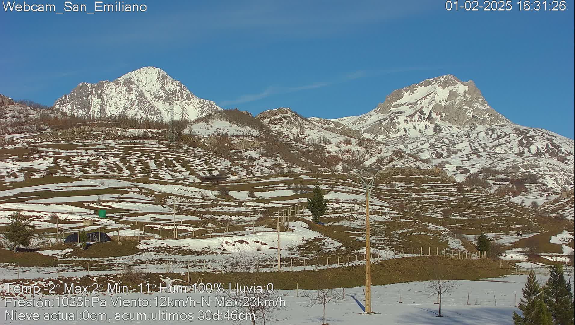 Two snow-capped mountains rise above a hilly landscape dotted with patches of snow under a clear blue sky.