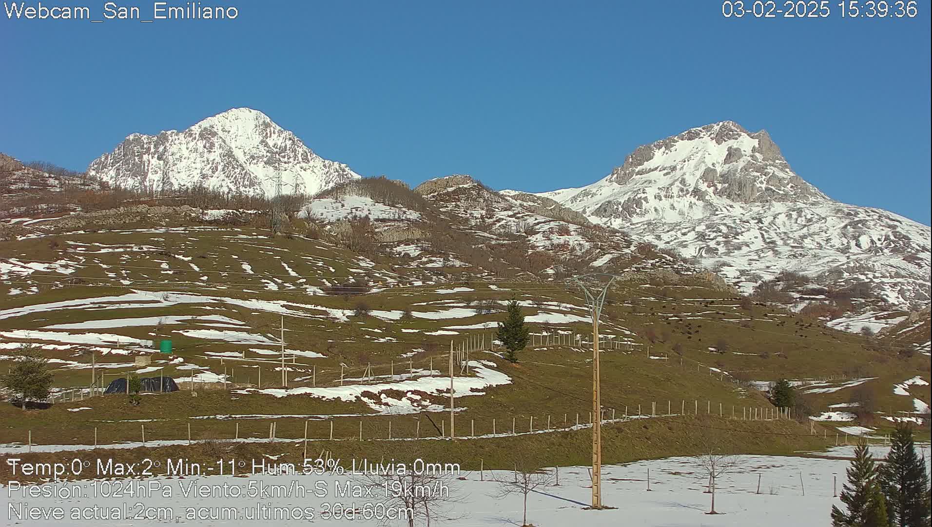 Two snow-capped mountains rise in the distance under a clear blue sky, overlooking a grassy valley with patches of snow.