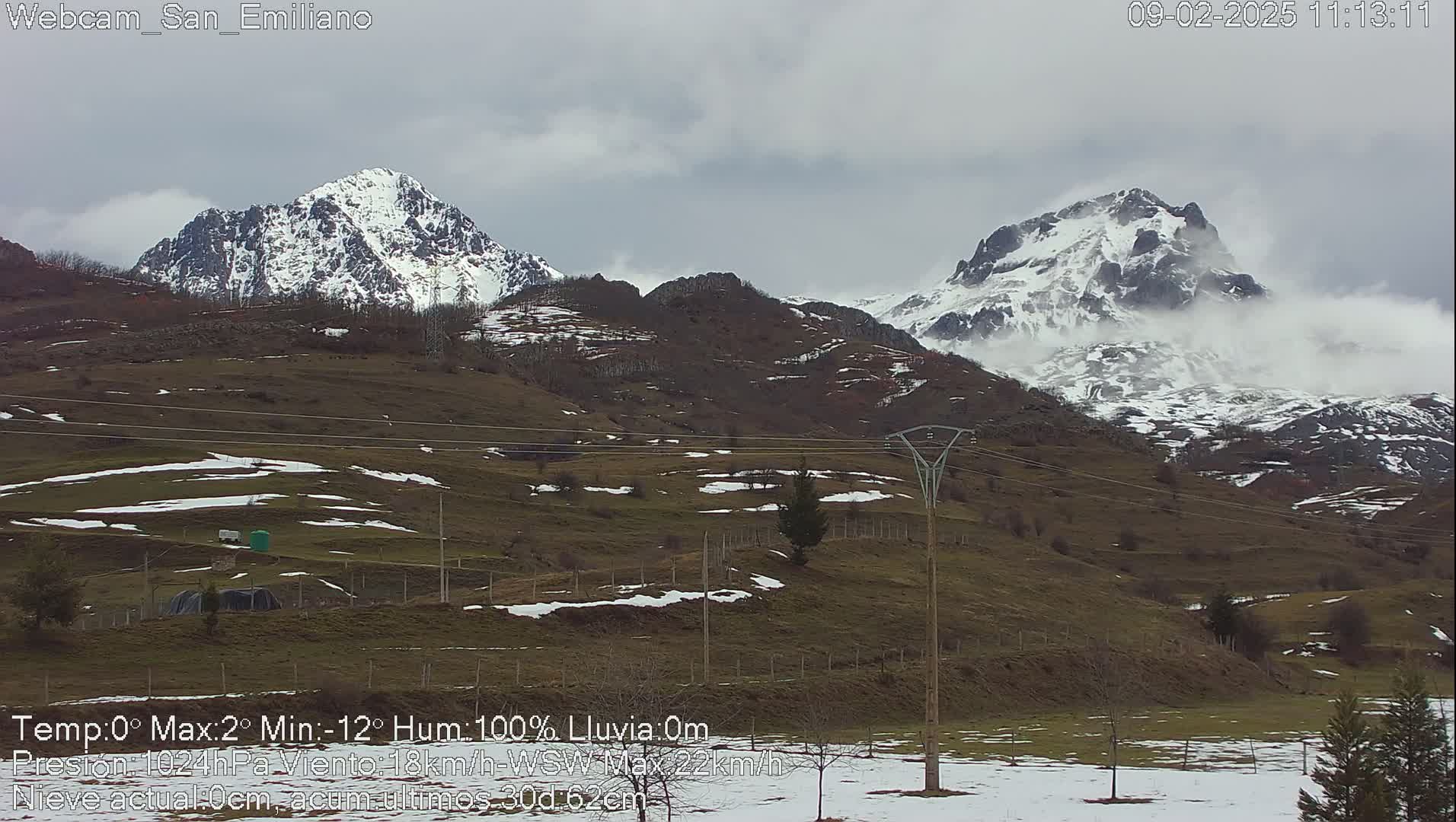 A view of snow-capped mountains under a cloudy sky, with patches of snow on the brown, grassy hills in the foreground.