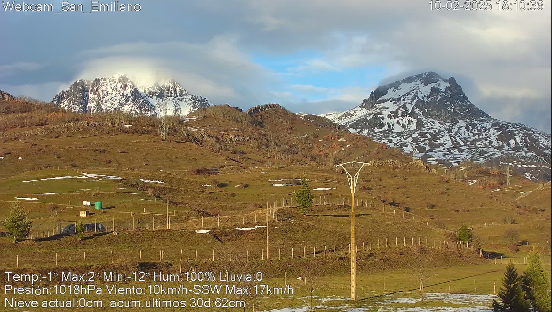 A partly cloudy day reveals snow-capped mountains in the distance, overlooking a grassy, fenced-in pasture with a few scattered trees and some small structures.