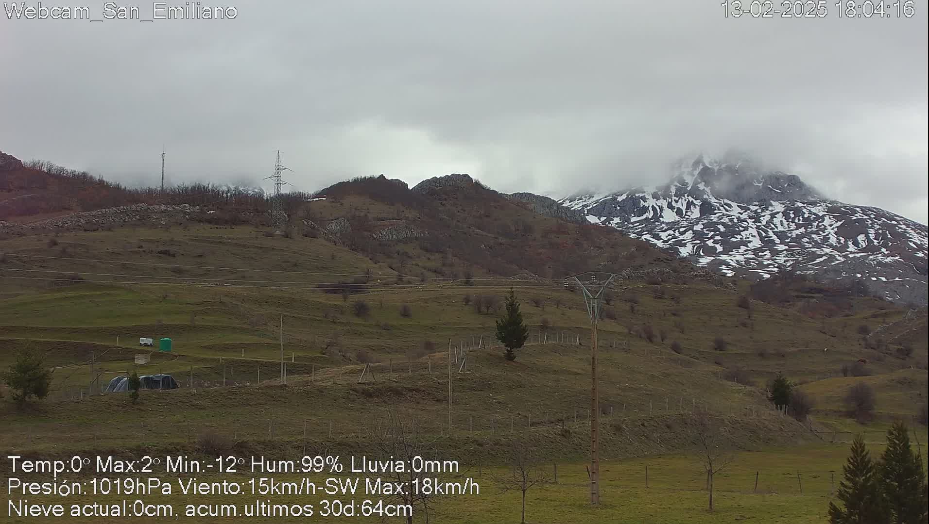 A snow-dusted mountain range is partially obscured by a cloudy sky over a grassy valley with sparse vegetation.