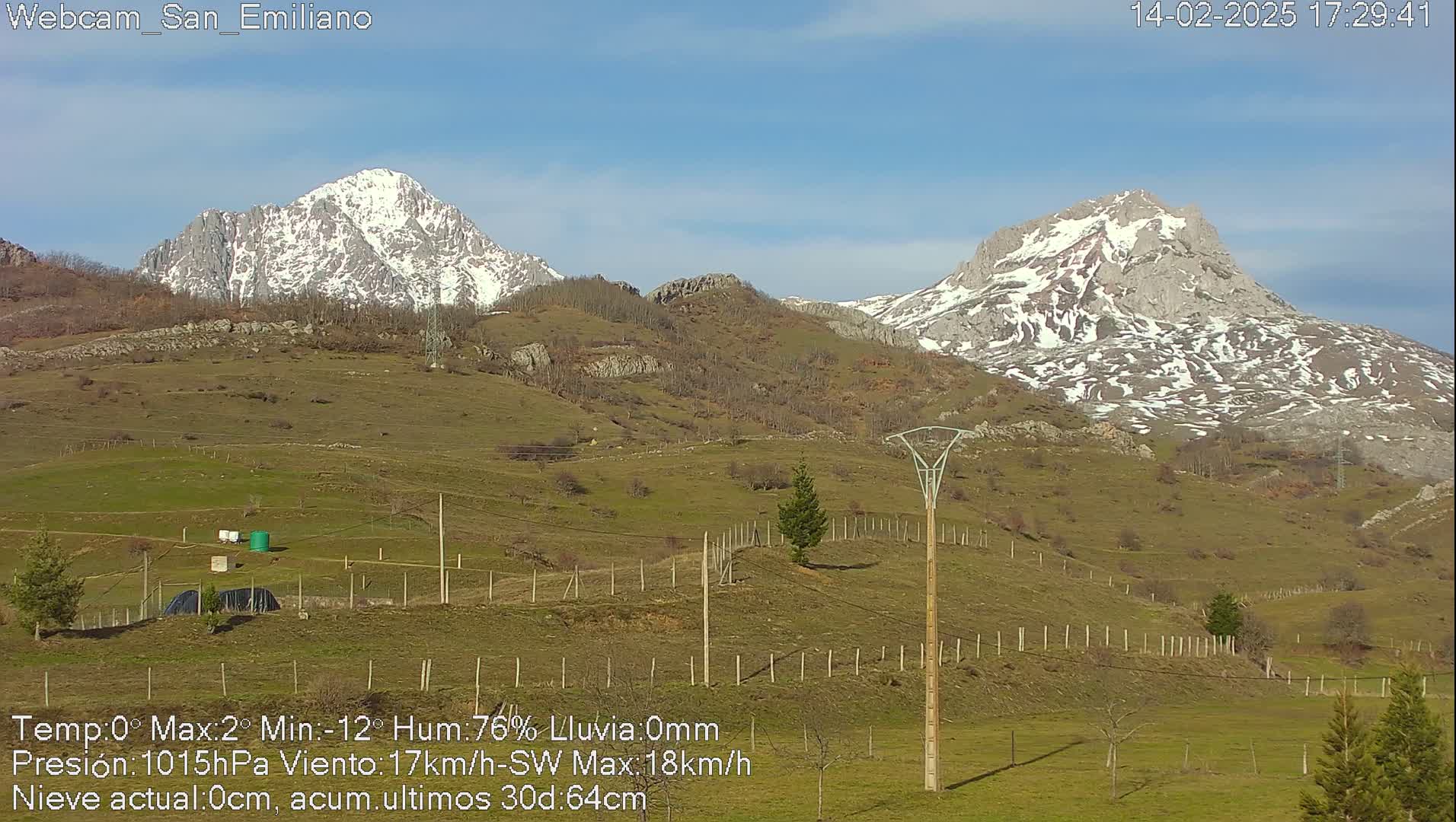 Two snow-capped mountains rise in the background of a sunny, grassy valley, with a few trees and utility poles in the foreground.