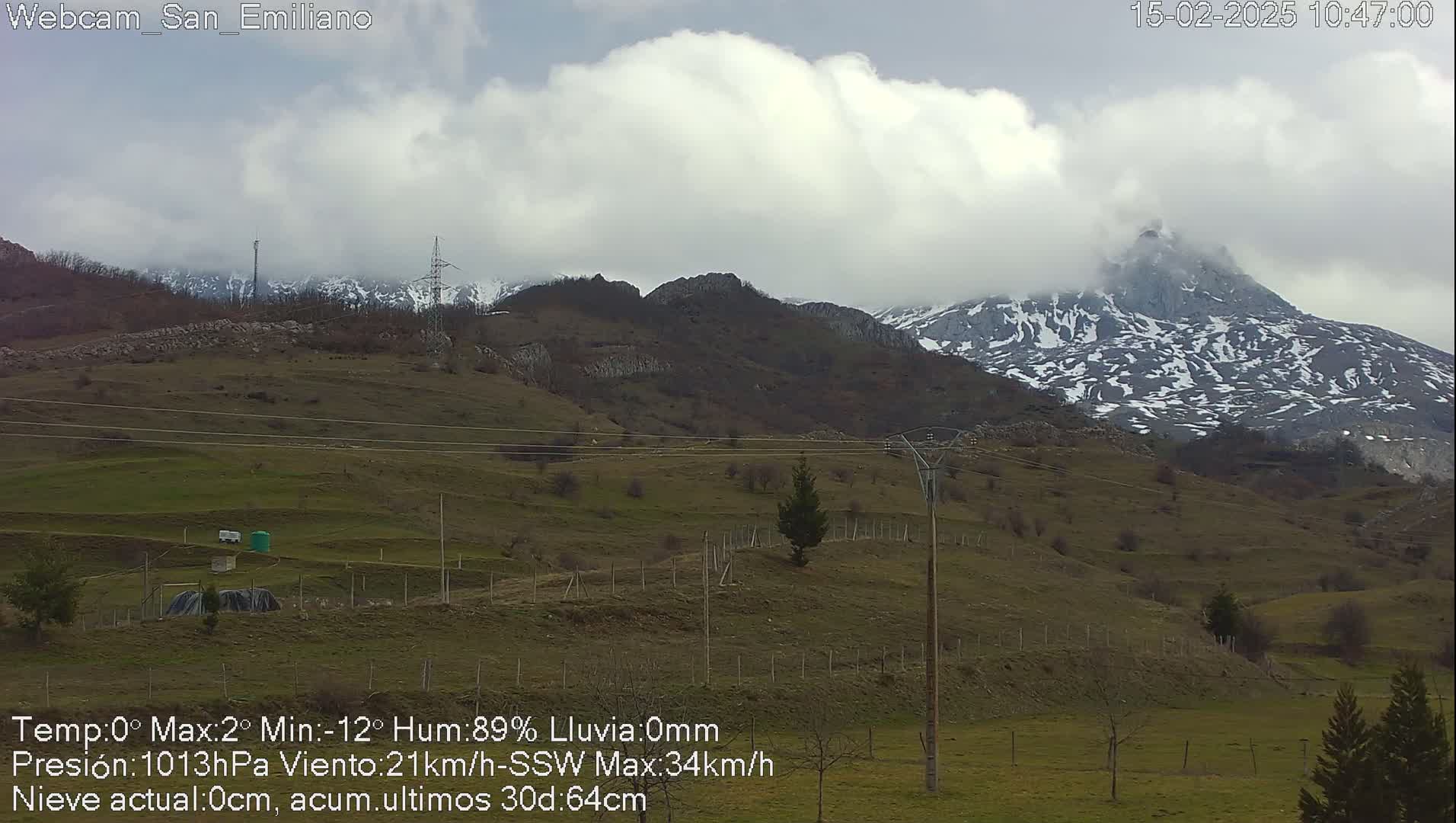 A partly cloudy day reveals a snow-dusted mountain range in the distance, overlooking a hilly, grassy landscape with sparse vegetation and power lines.