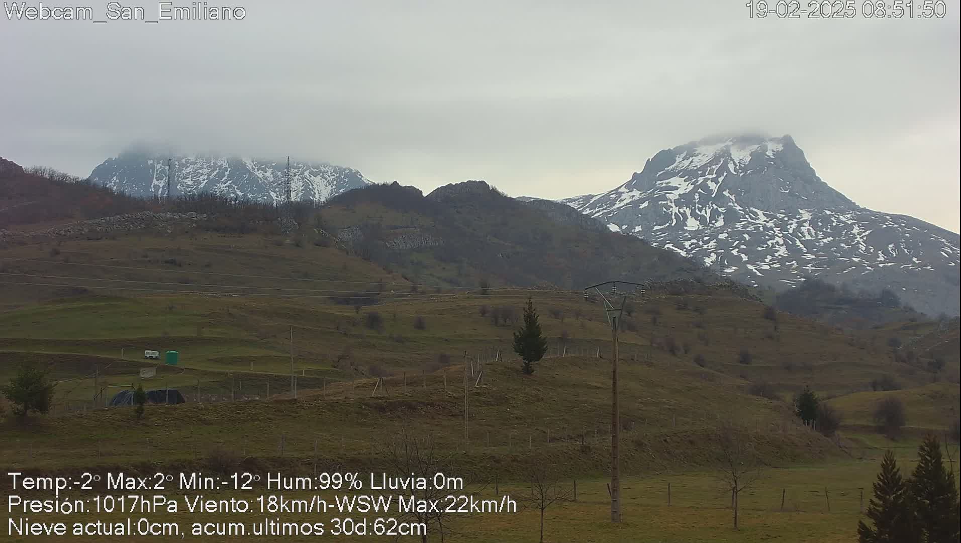 A view of snow-capped mountains under a cloudy sky, overlooking a sparsely vegetated hillside.