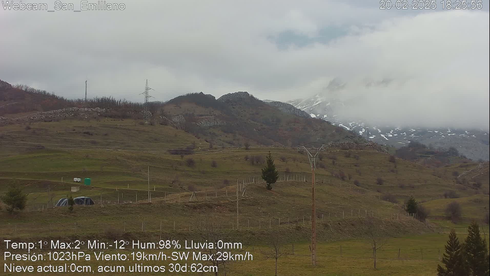 A hilly landscape with sparsely vegetated fields and distant snow-capped mountains under a cloudy, overcast sky.