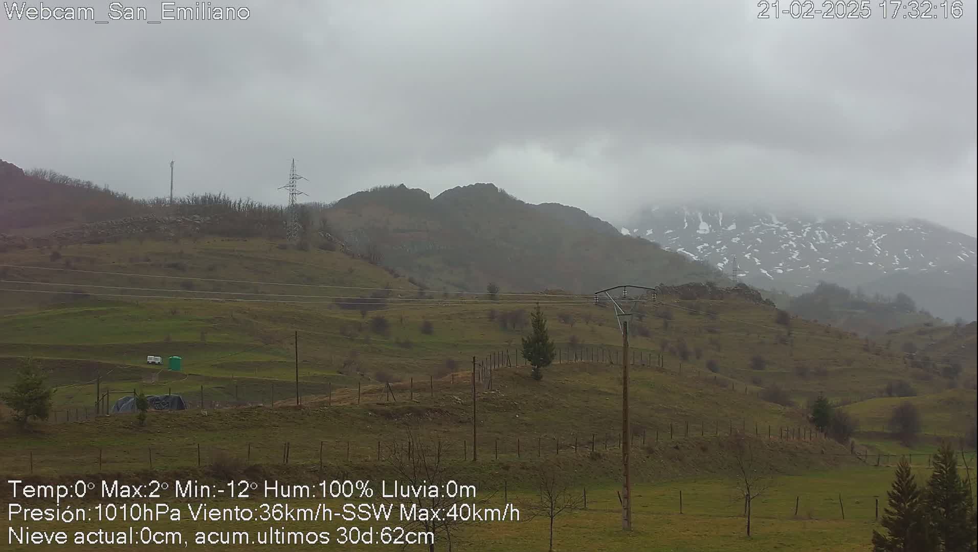 A foggy, overcast landscape shows rolling green hills and distant mountains with patches of snow under a gray sky.