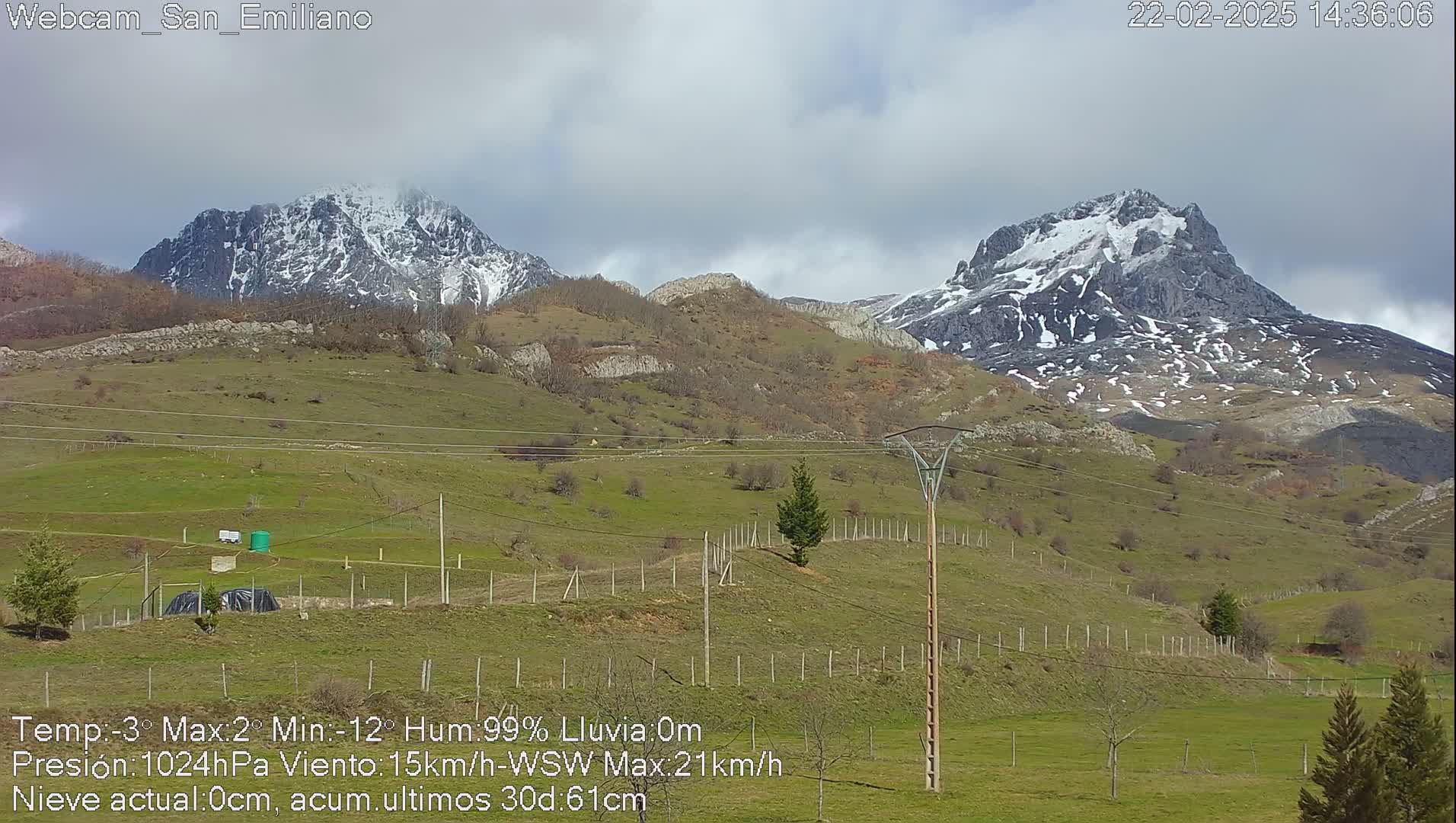 A partly cloudy day reveals snow-capped mountains in the distance, overlooking a grassy valley with a wooden fence and a lone utility pole.
