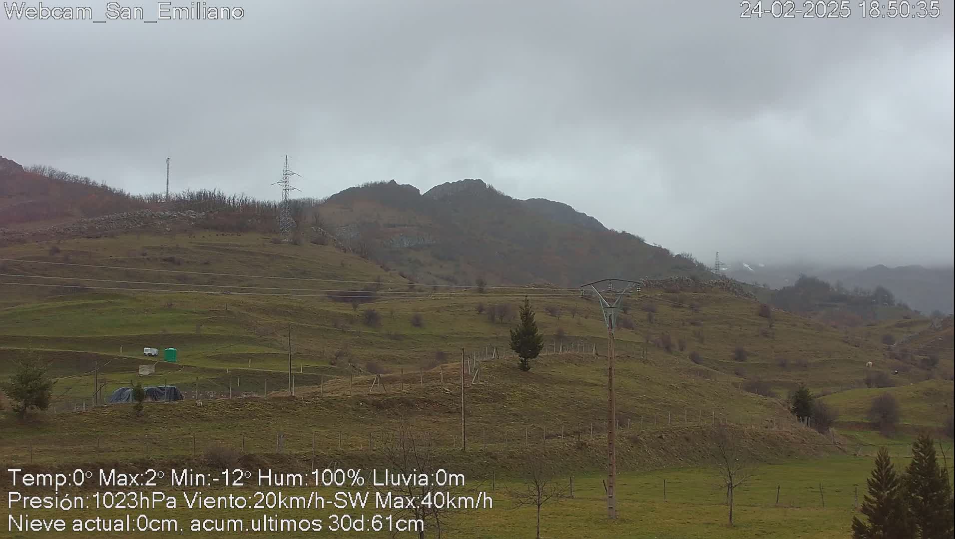 A hilly, sparsely vegetated landscape under an overcast sky is shown, with a few utility poles and some small structures visible in the foreground.