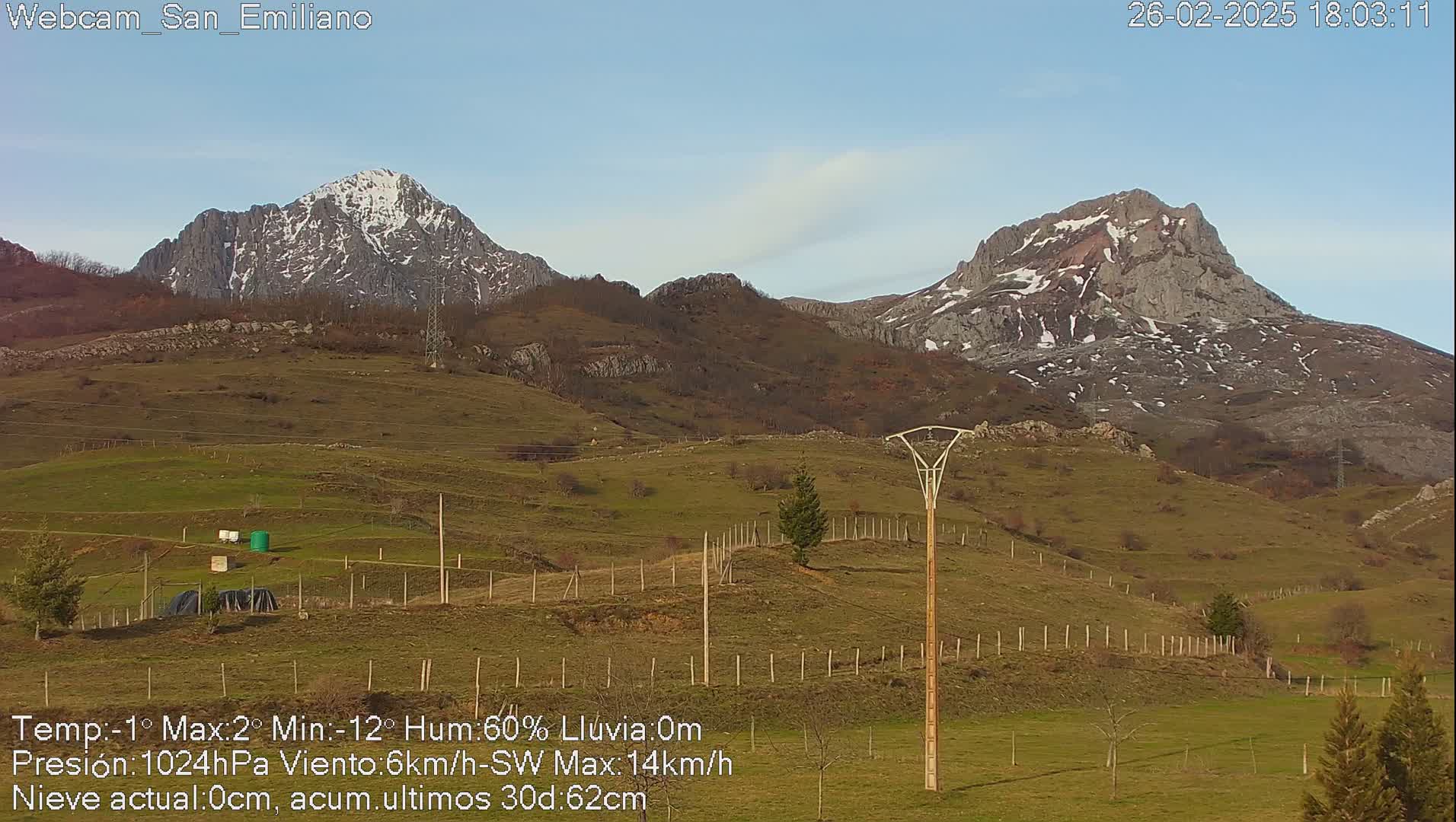 A clear, sunny view of snow-capped mountains and a grassy field with a wooden utility pole and fencing.