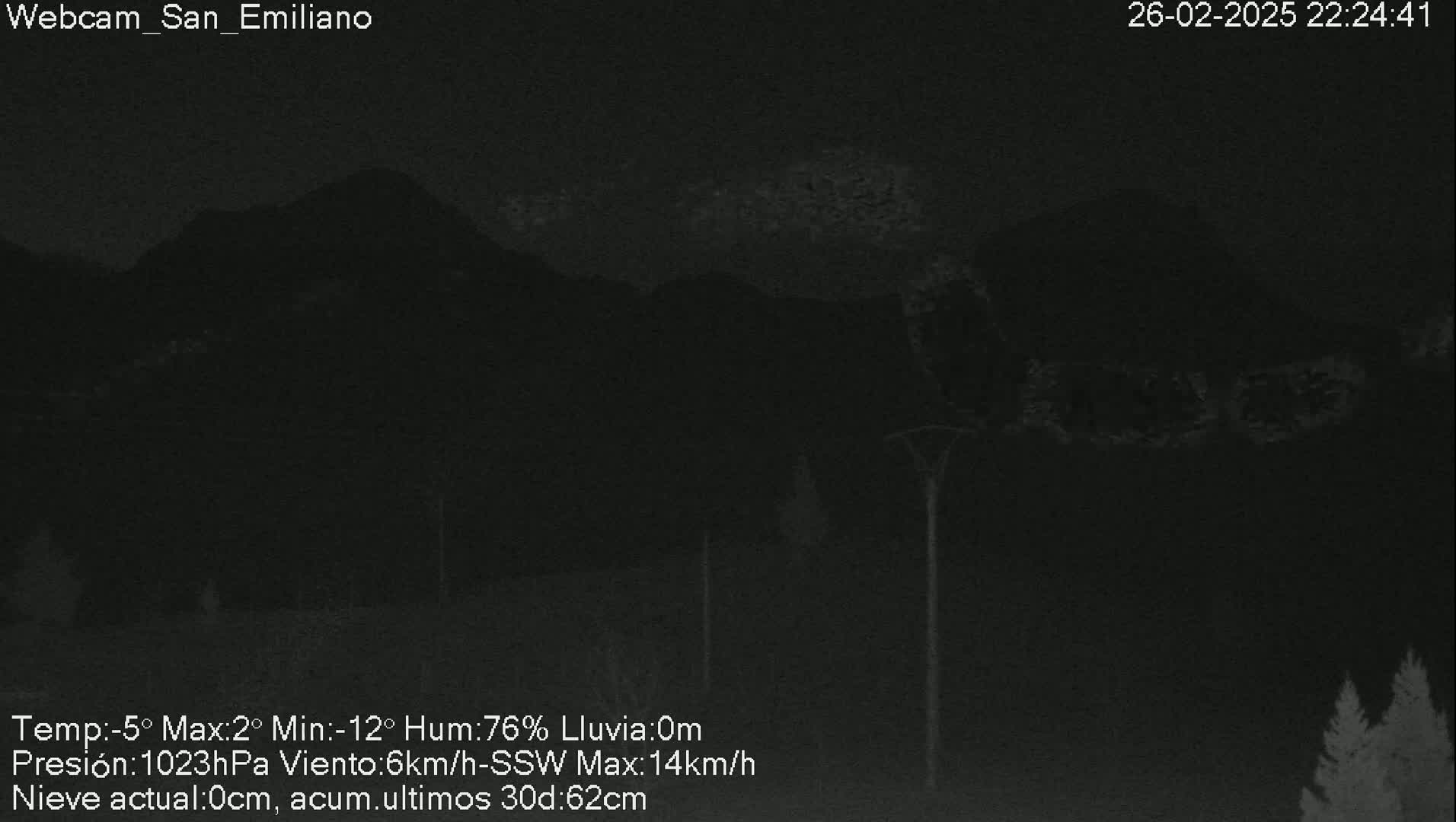 A nighttime, low-light image shows dark silhouettes of mountains under a cloudy sky, with some trees visible in the lower right corner and tall poles in the middle ground.