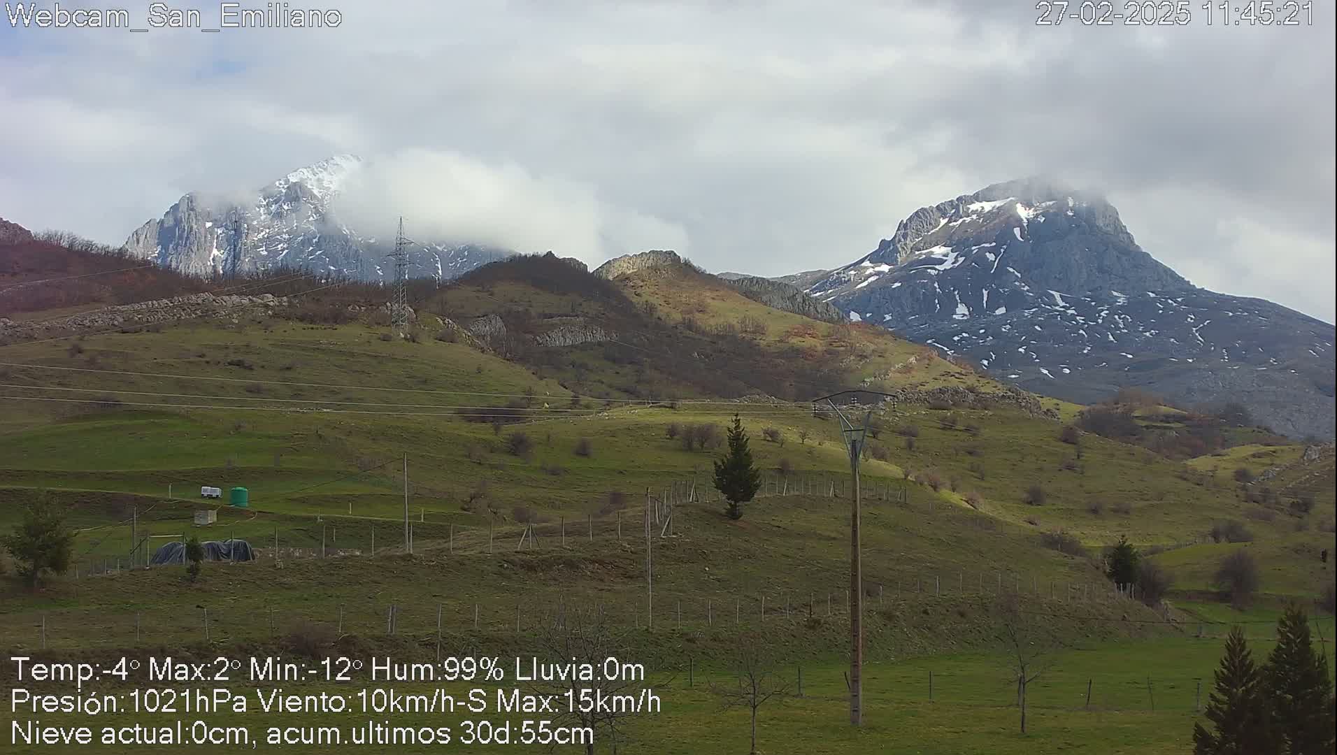 Snow-capped mountains rise above a green, hilly landscape under a cloudy sky.