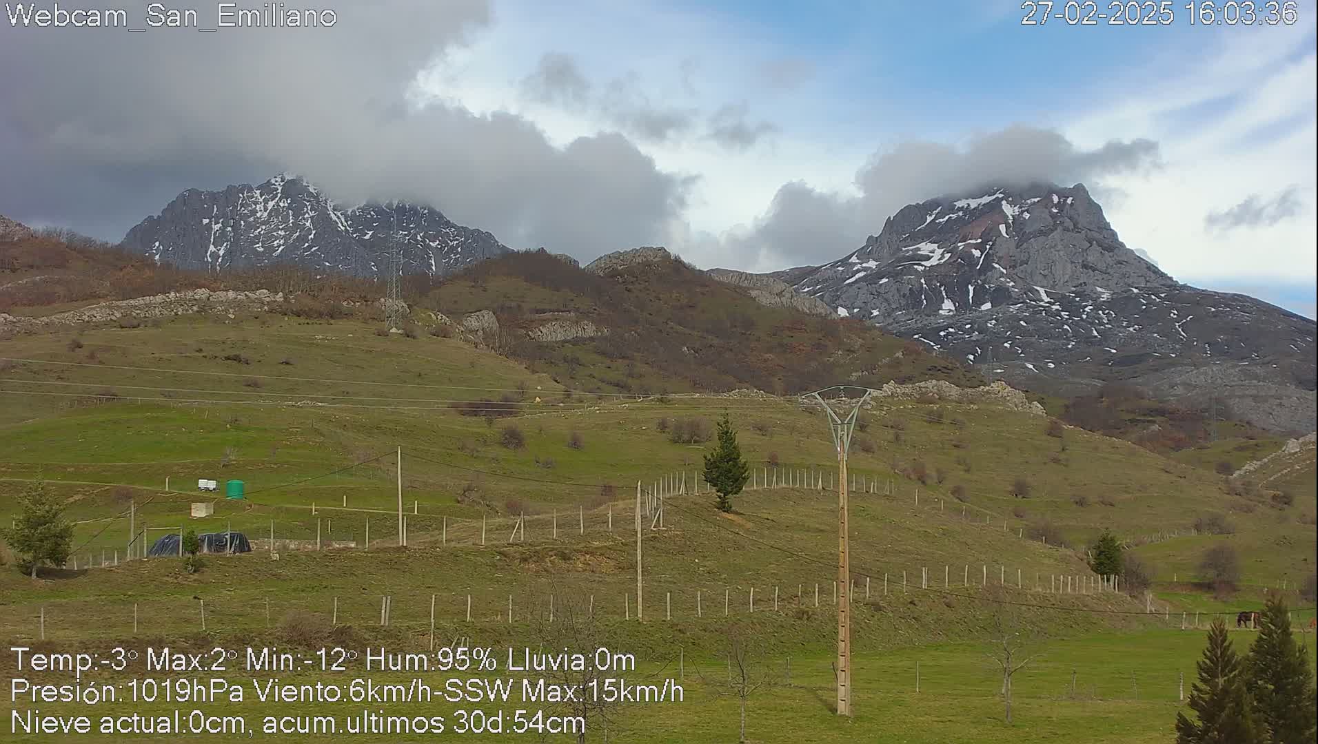 A partly cloudy day reveals snow-capped mountains in the distance, overlooking a grassy hillside with a wooden fence and utility poles.