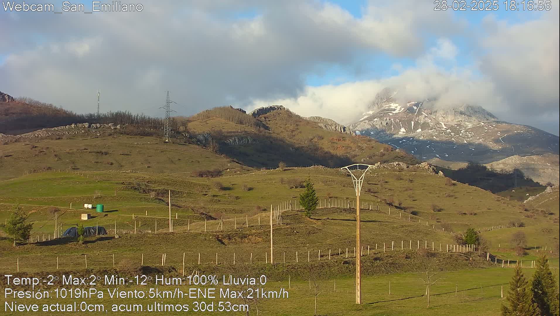 A partly cloudy day reveals a snow-capped mountain range in the background, overlooking a grassy hillside dotted with fences and utility poles.