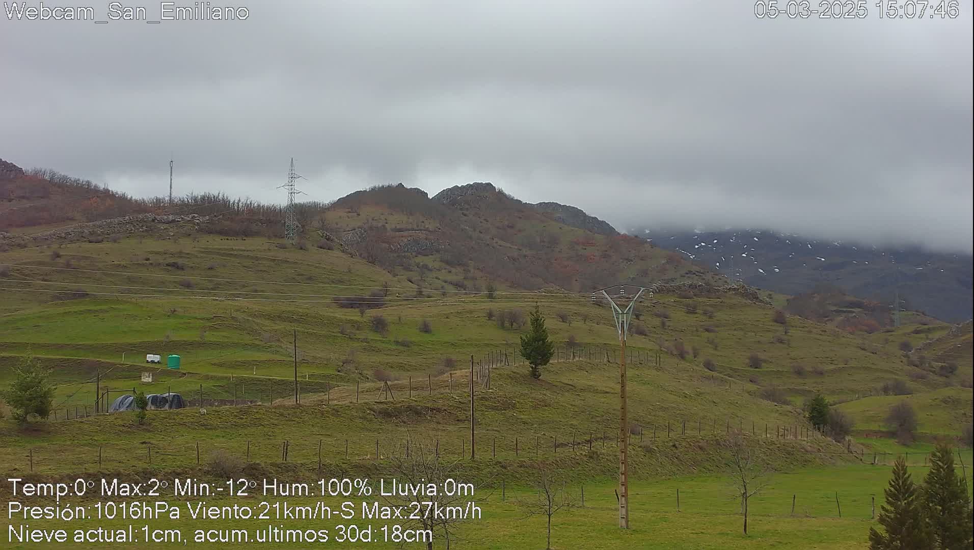 A hilly, green landscape under a cloudy, overcast sky shows some snow on distant mountains.