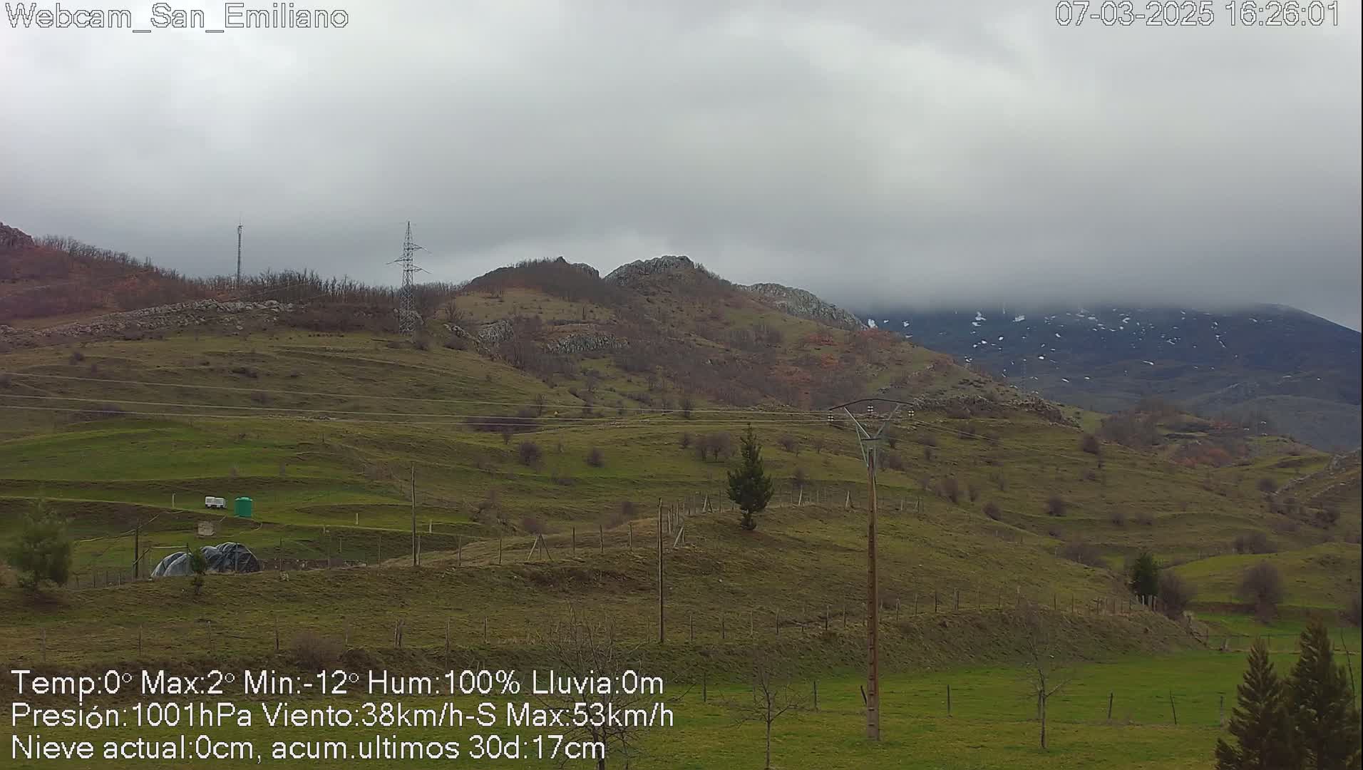 A view of rolling green hills under a cloudy, overcast sky, with distant mountains partially covered in snow.