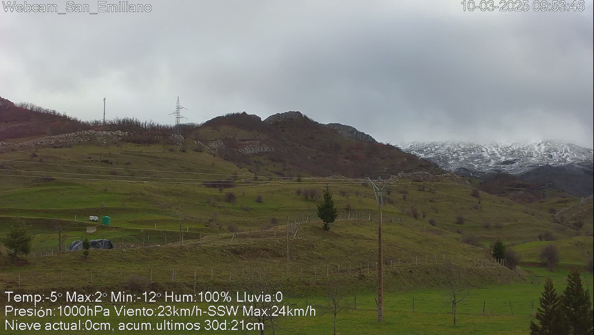 A snow-dusted mountain range is visible in the distance under an overcast sky, with green rolling hills and sparse vegetation in the foreground.