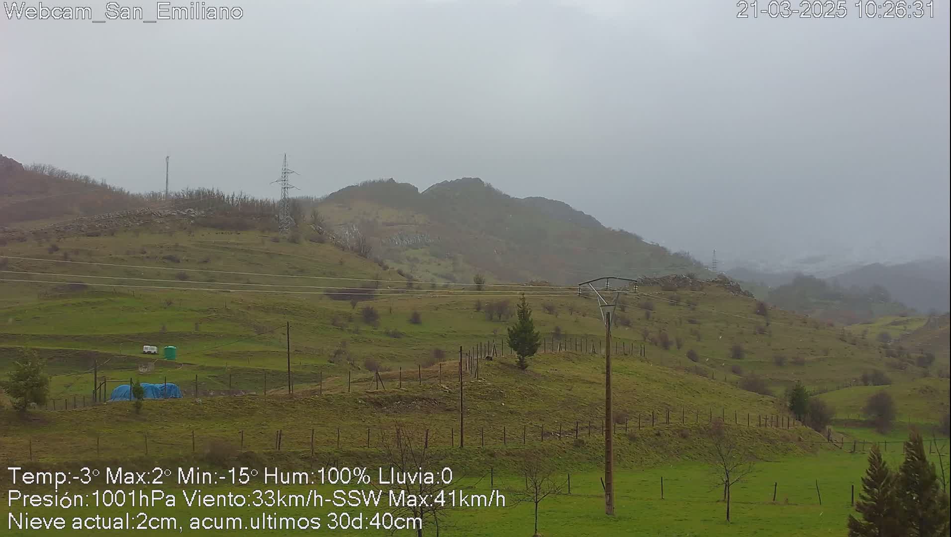 A misty, overcast view of rolling green hills and a few sparsely wooded areas, with power lines traversing the landscape.