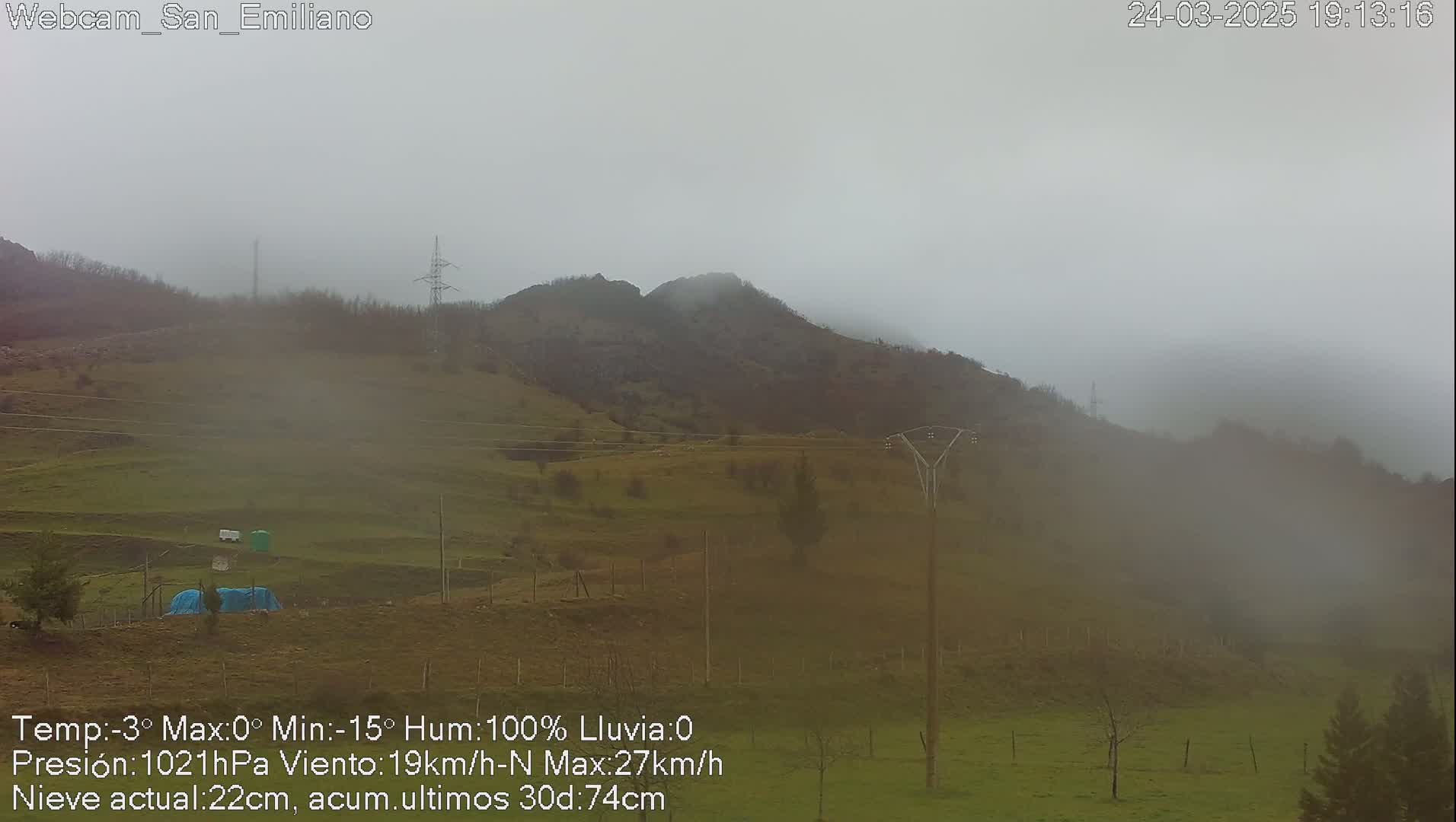A foggy, overcast scene shows a hillside with sparse vegetation, power lines, and a few small structures in the foreground.