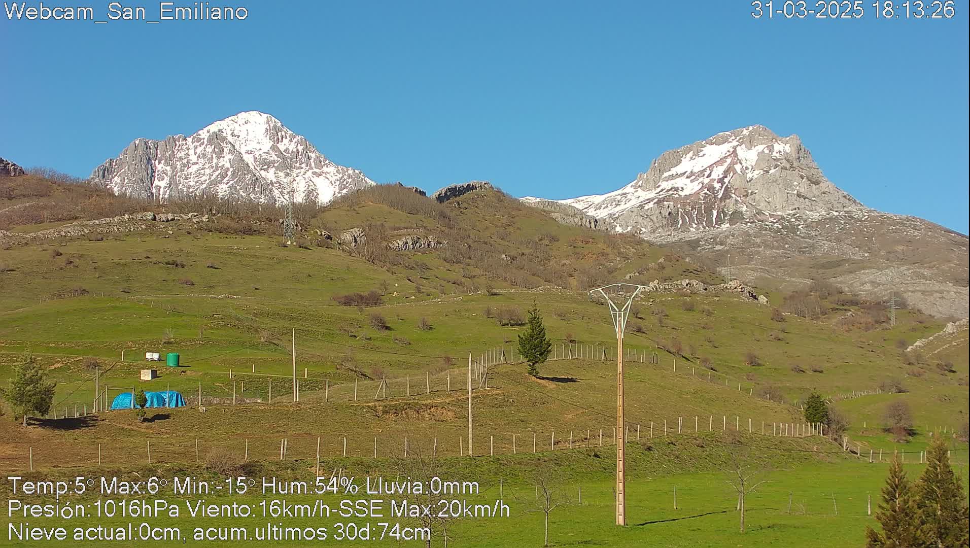 A sunny, snow-capped mountain range is visible in the distance from a grassy hillside with some fences and a few small structures.