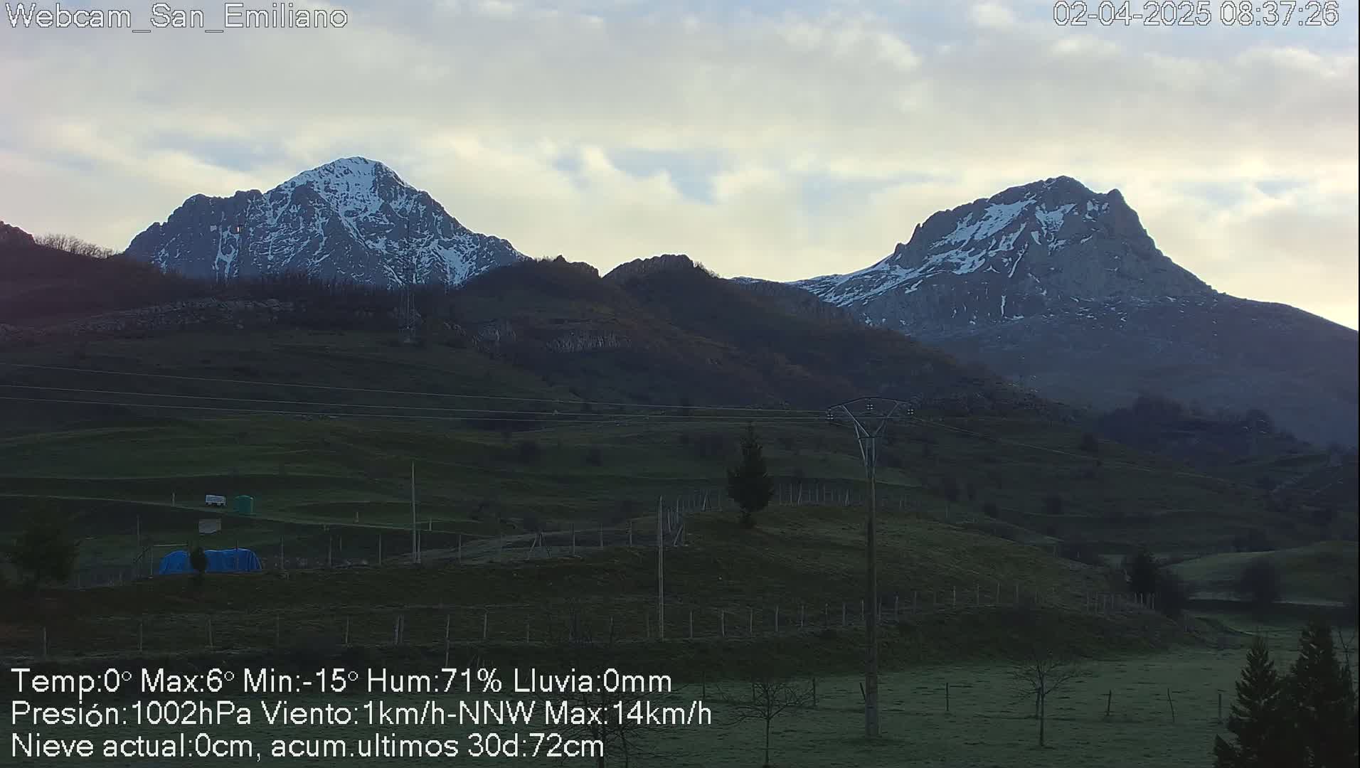 Two snow-capped mountains rise in the distance over a gently sloping, green valley under a mostly clear, light grey sky.