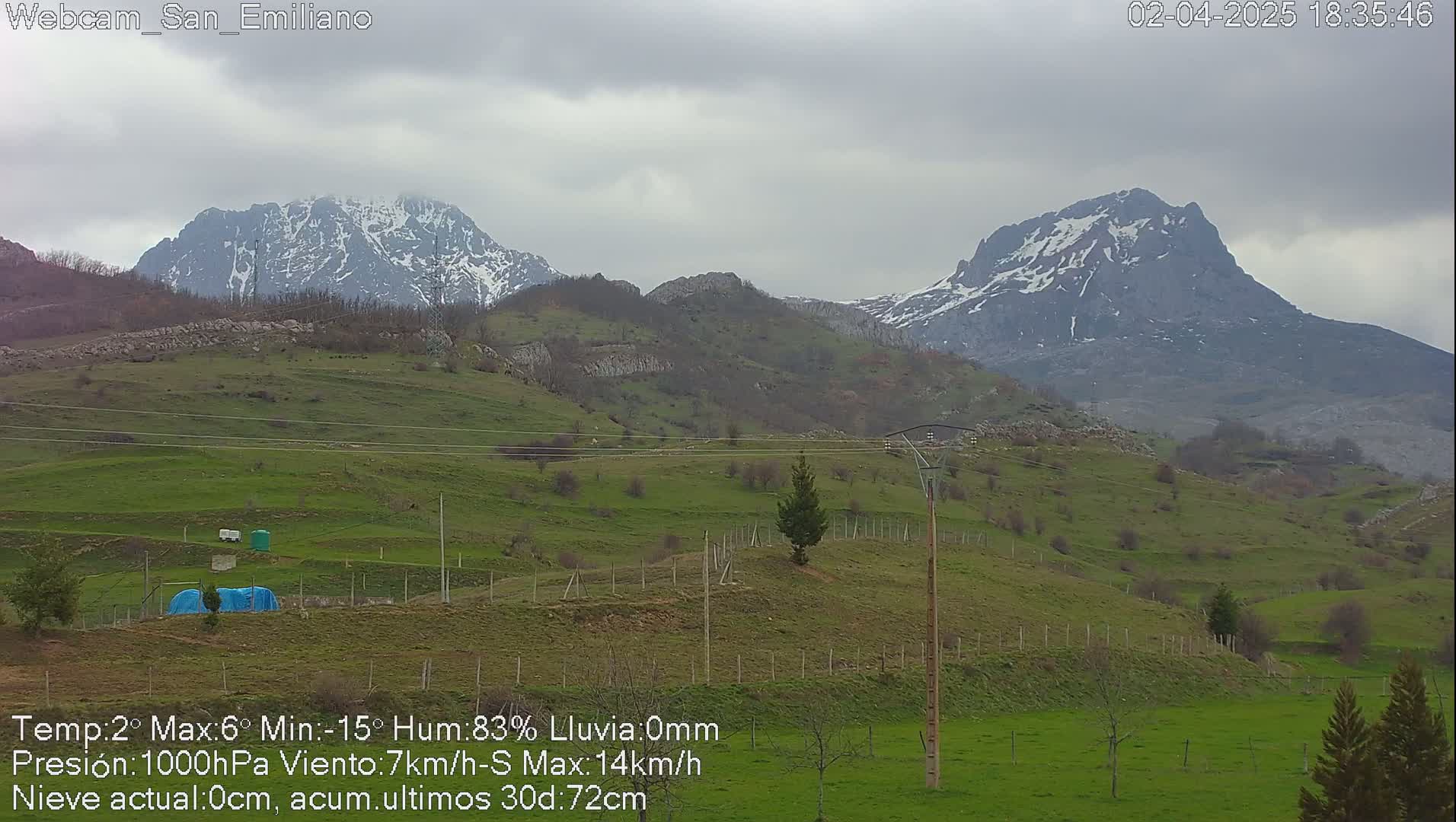 Snow-capped mountains rise in the distance over a gently rolling landscape of green hills and sparse trees under an overcast sky.