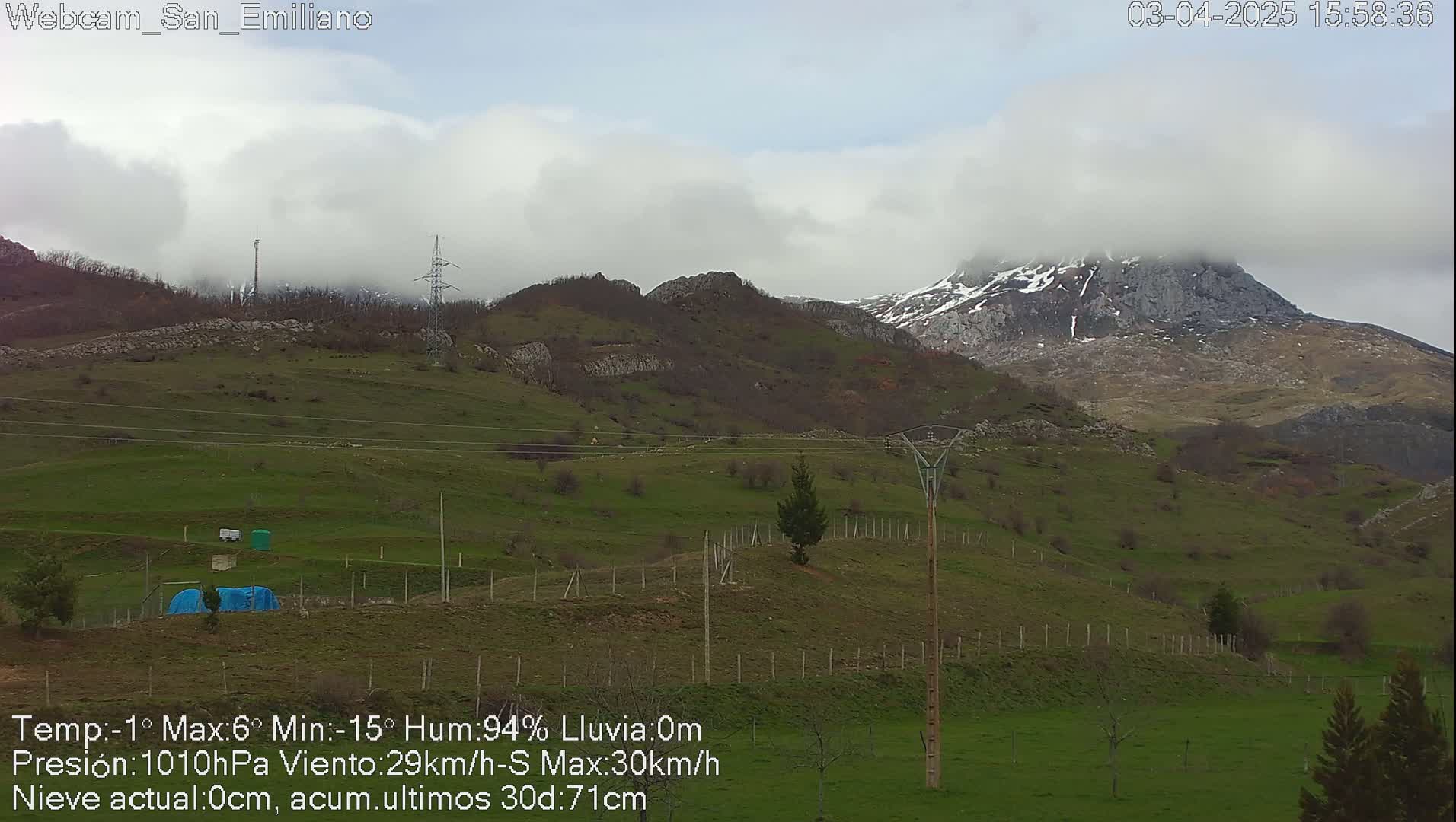 A partly cloudy, windy day shows a green, hilly landscape with a snow-capped mountain in the background.