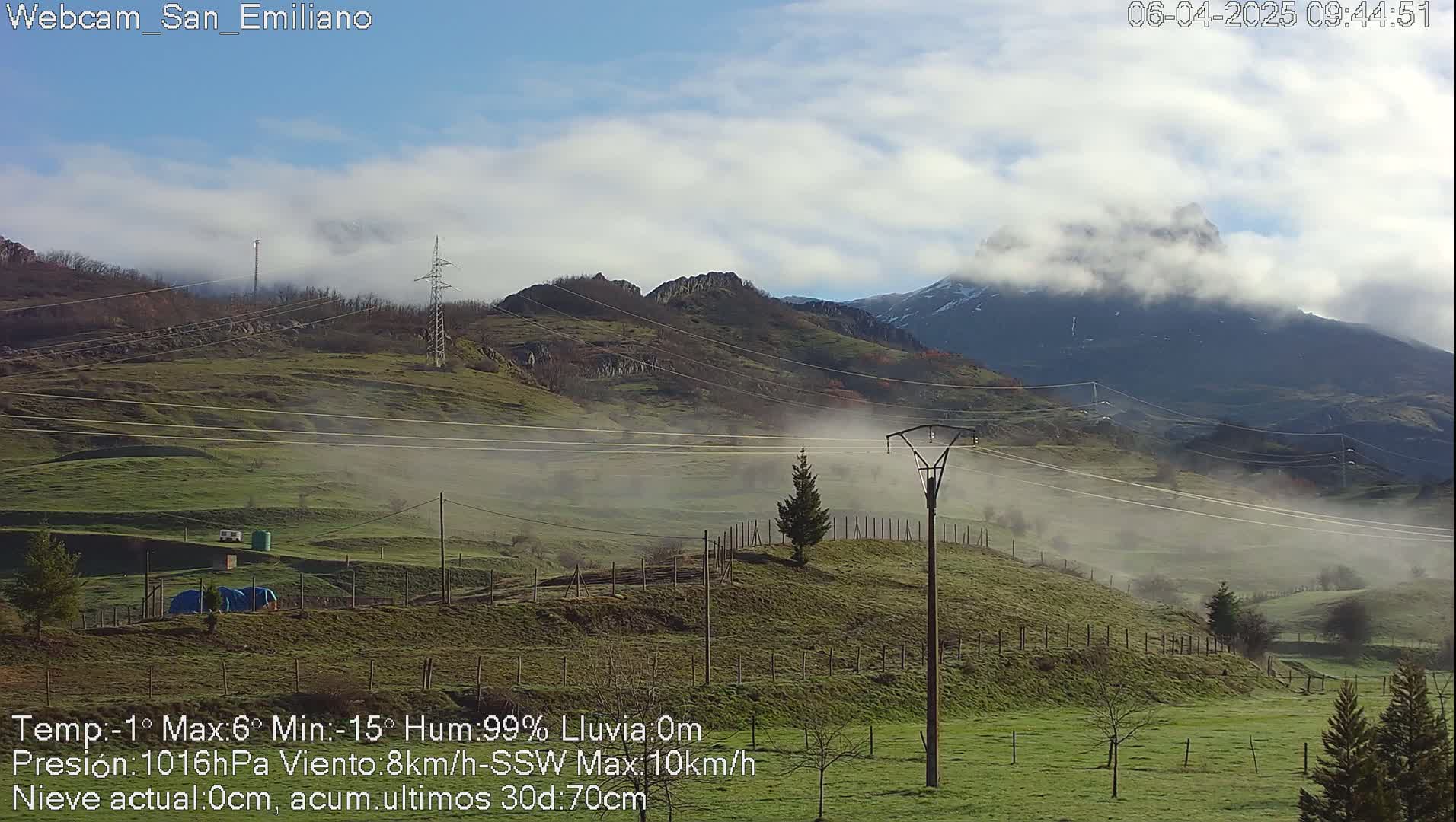 A misty, partly cloudy landscape shows rolling green hills, power lines, and a distant mountain range with a peak partially obscured by clouds.