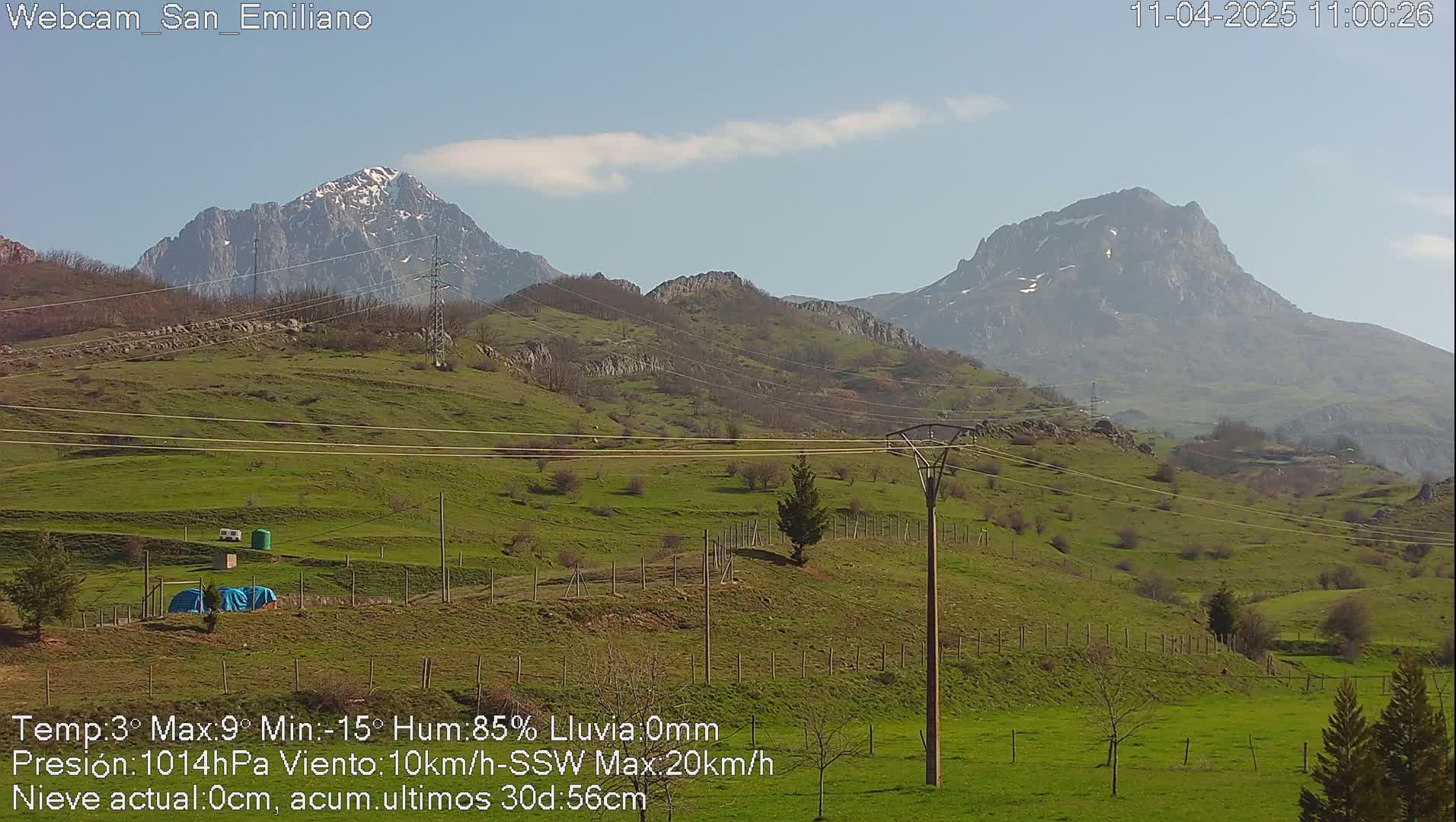 A sunny, green valley with power lines stretches between two snow-capped mountains under a mostly clear sky.