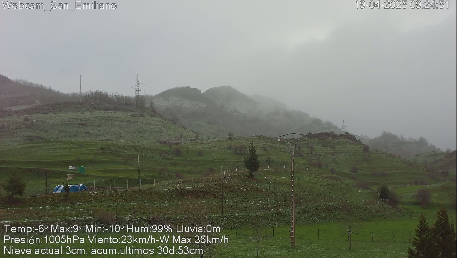 A misty, overcast scene shows rolling green hills with some light snow dusting the ground under a gray sky.