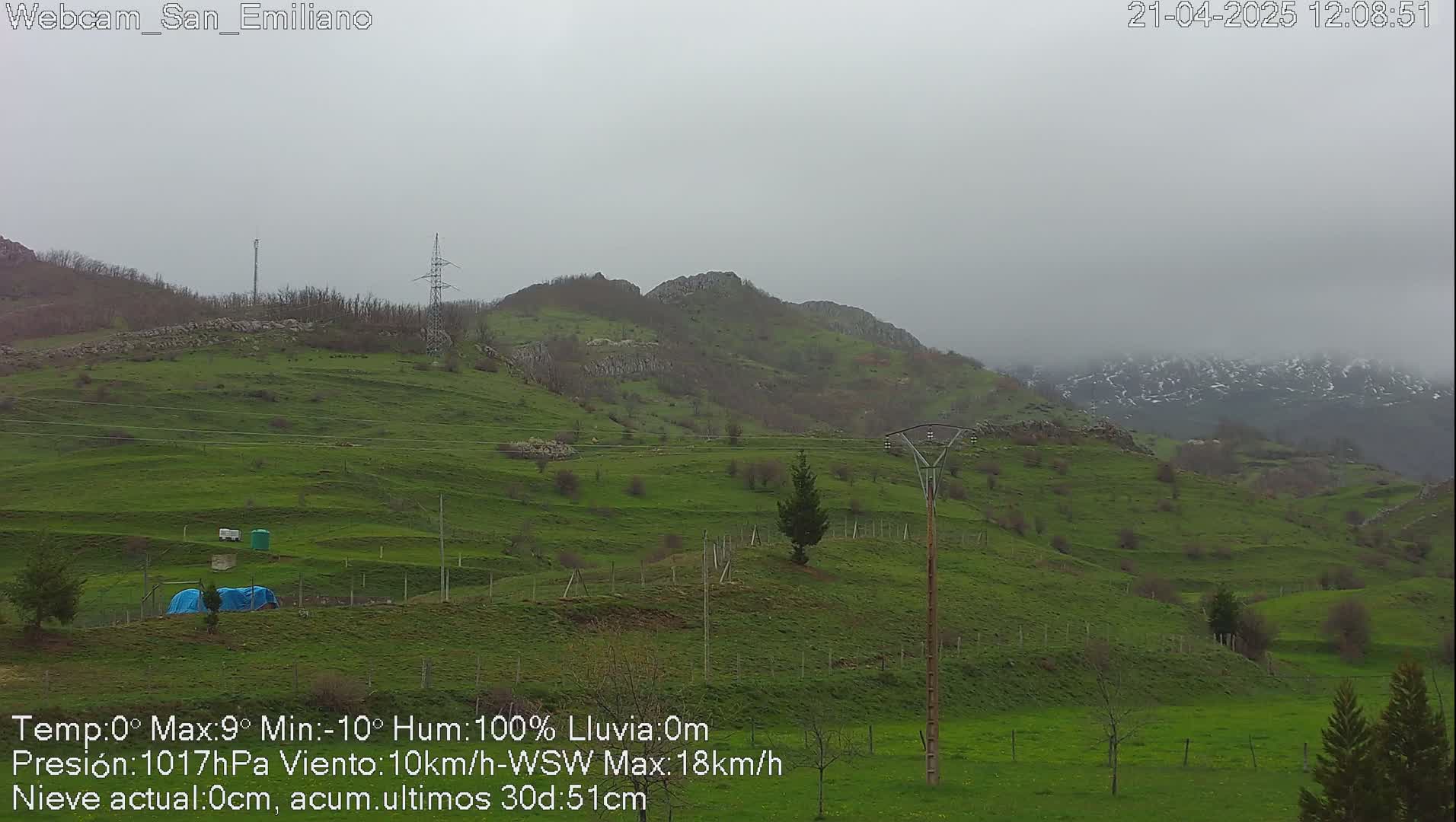 A misty, overcast view of rolling green hills and distant mountains, some with patches of snow, under a gray sky.