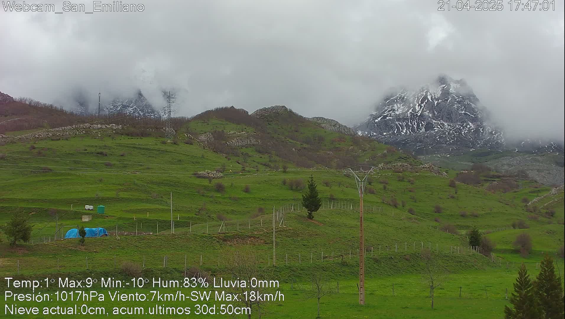 A mostly cloudy, slightly foggy scene shows a snow-dusted mountain peak rising above rolling green hills and farmland.